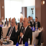 Businessman in a suit speaks into a microphone at a round table conference with attendees listening in a bright formal meeting room   جامعة الرافدين   مدير الموقع عمار محمود ابو الشعير   جامعة الرافدين   مدير الموقع عمار محمود ابو الشعير