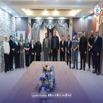 Group of officials in formal attire posing behind a long flowered table at a ceremonial event in a conference room.