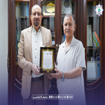 Two men in a formal setting exchange a framed certificate; they stand before bookshelves.