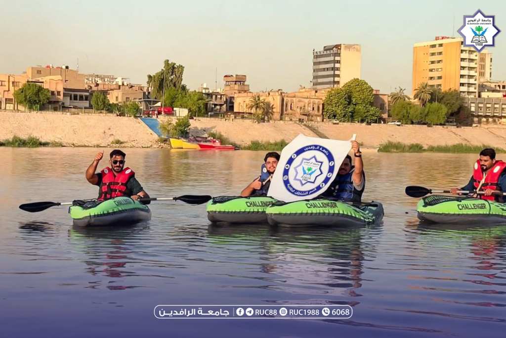 WhatsApp Image 20260831 at 114323 AM 1 جامعة الرافدين 2026 Three people in bright green inflatable kayaks wearing life jackets paddling on a calm river one person holds up a circular banner with a blue emblem amid an urban shoreline backdrop جامعة الرافدين مدير الموقع عمار محمود ابو الشعير