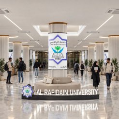 A spacious university lobby with a central pillar display reading 'Al Rafidain University' and students walking or sitting nearby.