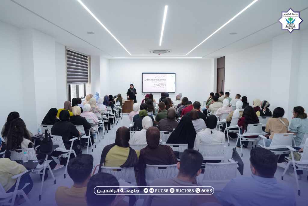 DSC03393 1 جامعة الرافدين 2026 University lecture hall a female speaker at a podium presents to a large audience with slides on screen and a university logo in the top right corner جامعة الرافدين مدير الموقع عمار محمود ابو الشعير