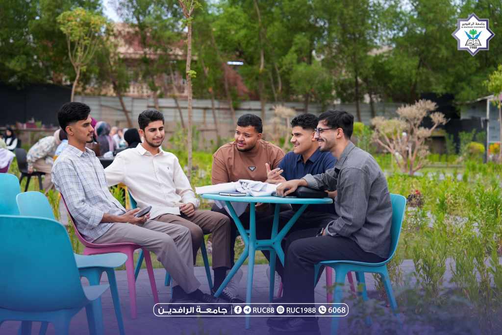 Group of five young men sitting around a blue table outdoors chatting in a university courtyard with trees and a campus logo in the top right corner   جامعة الرافدين   مدير الموقع عمار محمود ابو الشعير