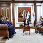 Formal meeting room with officials seated in two rows a man stands at a small table with a flag in the background   جامعة الرافدين   مدير الموقع عمار محمود ابو الشعير   جامعة الرافدين   مدير الموقع عمار محمود ابو الشعير